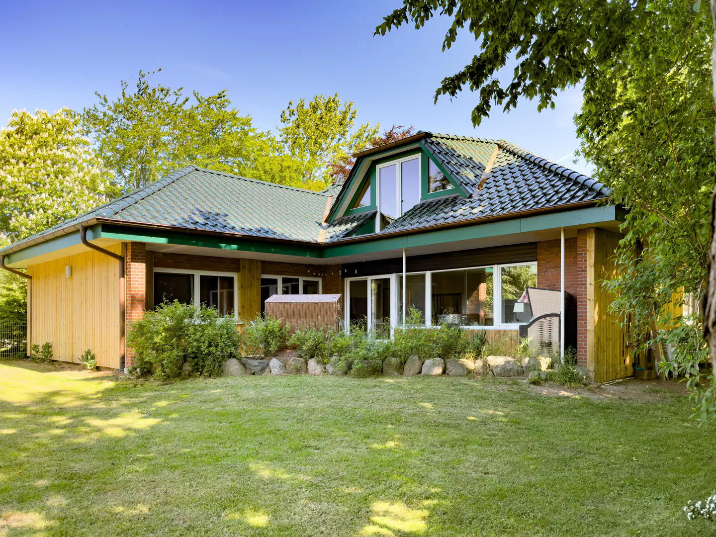 A modern house with a green tiled roof, brick and wood siding, and large windows overlooking a lush green lawn and trees.