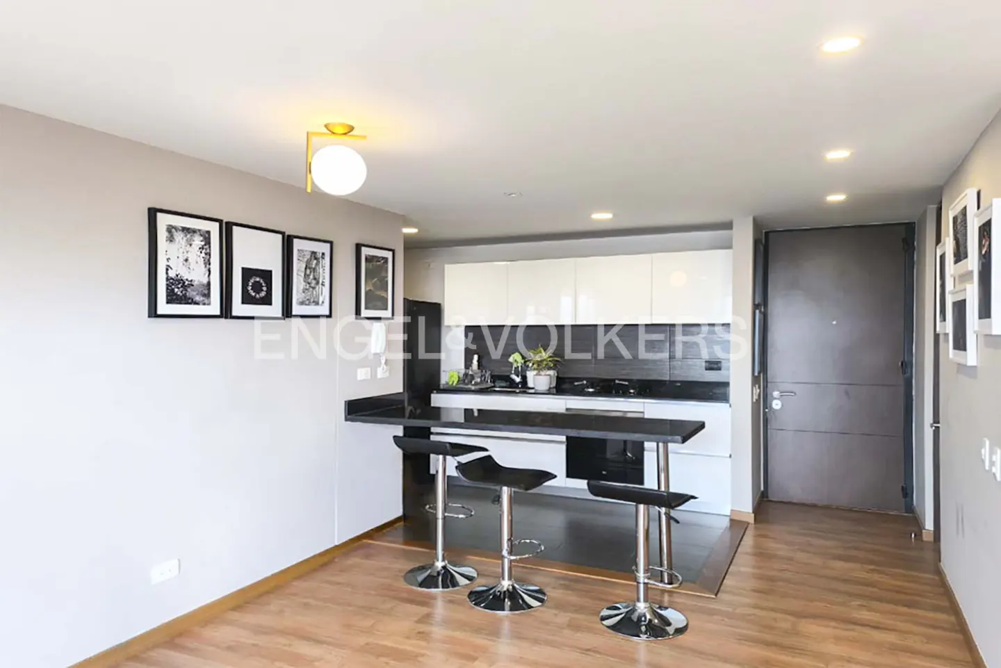 A modern apartment interior with a black countertop, three stools, and white cabinets. Artwork adorns the gray walls.