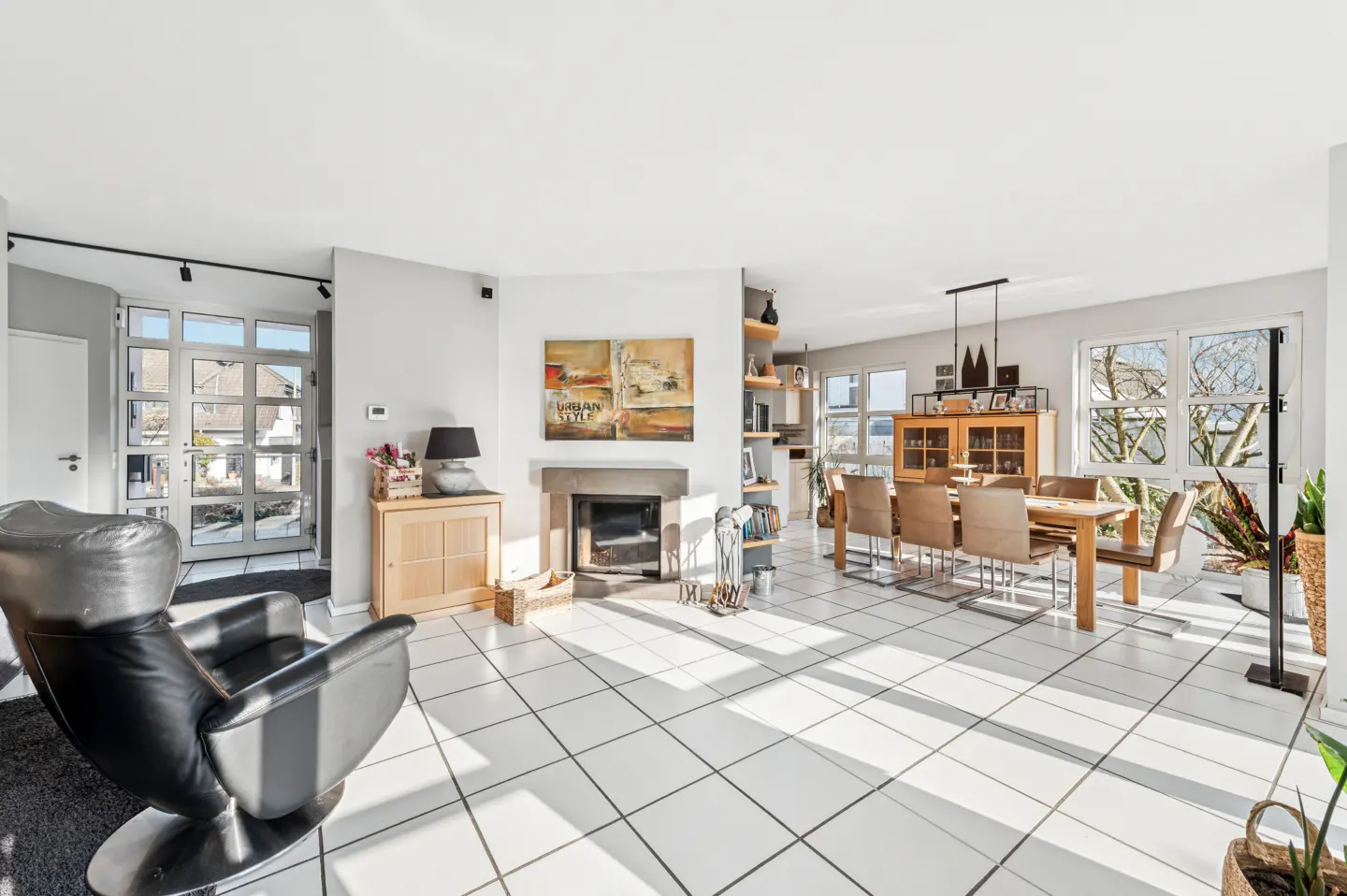 Bright, open-concept living and dining area with white tile floors. A black leather chair sits near a fireplace and dining table with six chairs.