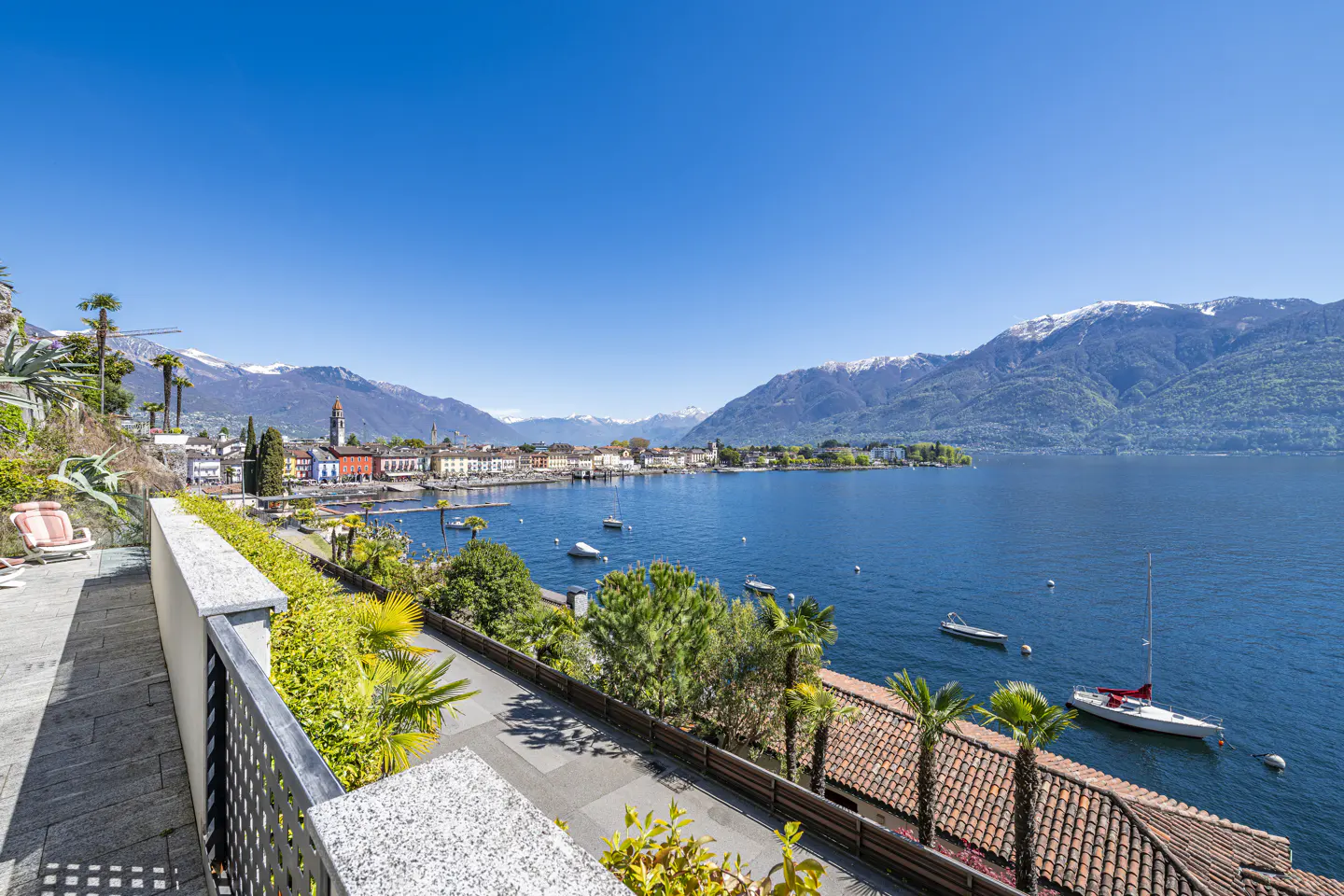 Scenic view of Lake Maggiore, Switzerland, with boats on blue water, mountains in the background, and a tiled roof in the foreground.