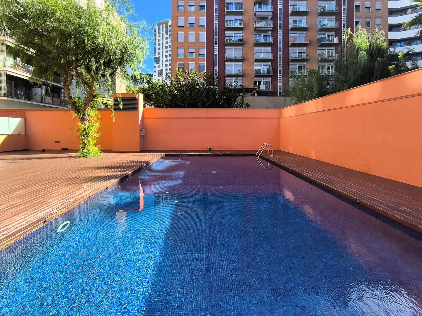 Outdoor pool with blue tile, surrounded by a wooden deck and orange walls. Tall buildings are visible in the background.