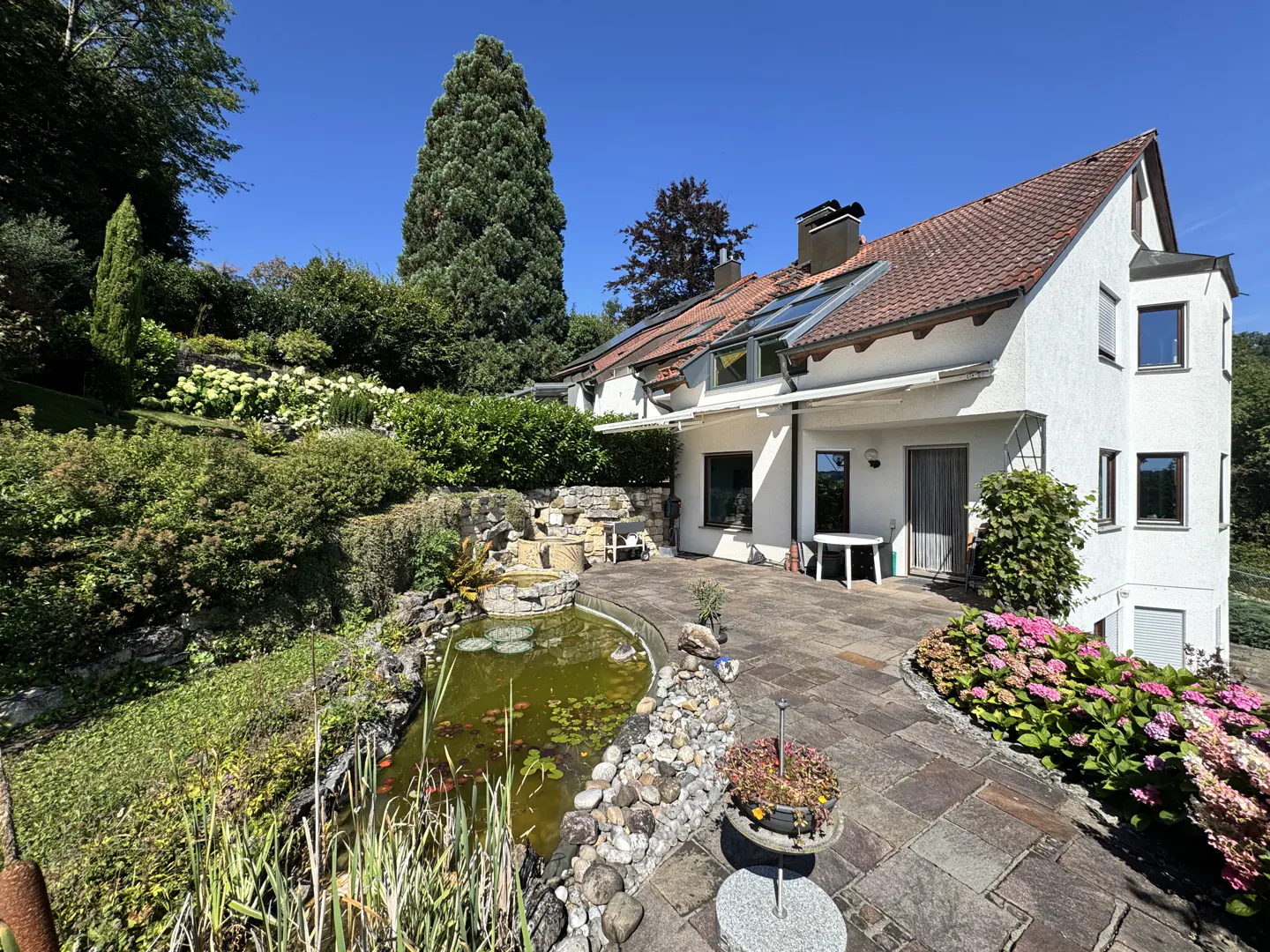 Exterior view of a white house with a red tile roof, a stone patio, and a pond with lily pads. Lush greenery surrounds the property.