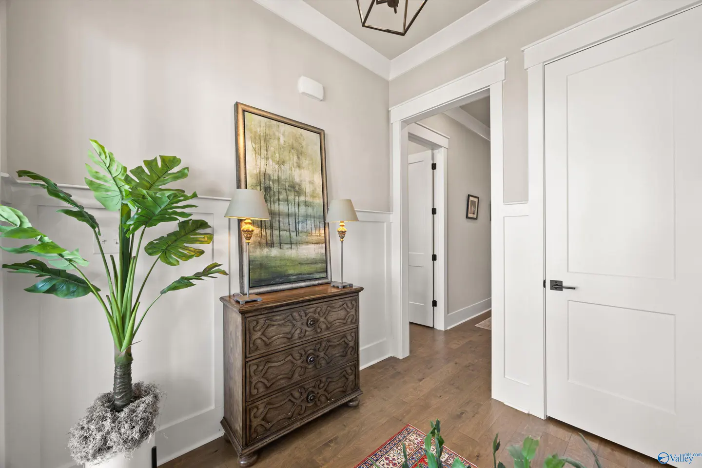 Foyer with hardwood floors, white walls, and trim. A carved wood dresser holds lamps and a framed landscape painting. A large green plant sits nearby.