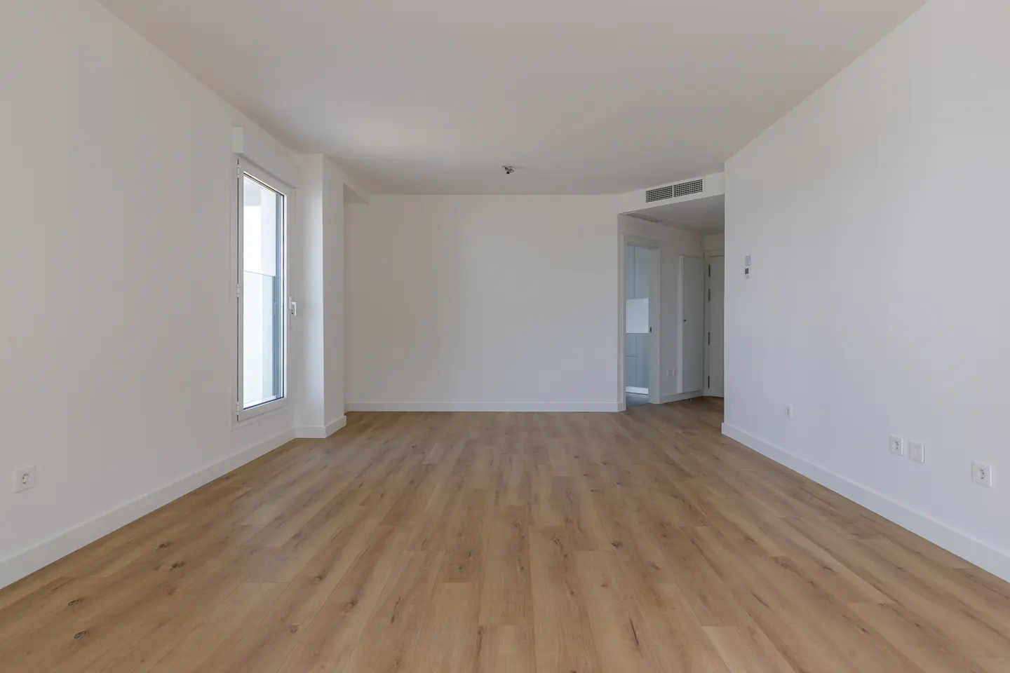 An empty room with white walls and light brown wood floors. A window is on the left, and a doorway is on the right.
