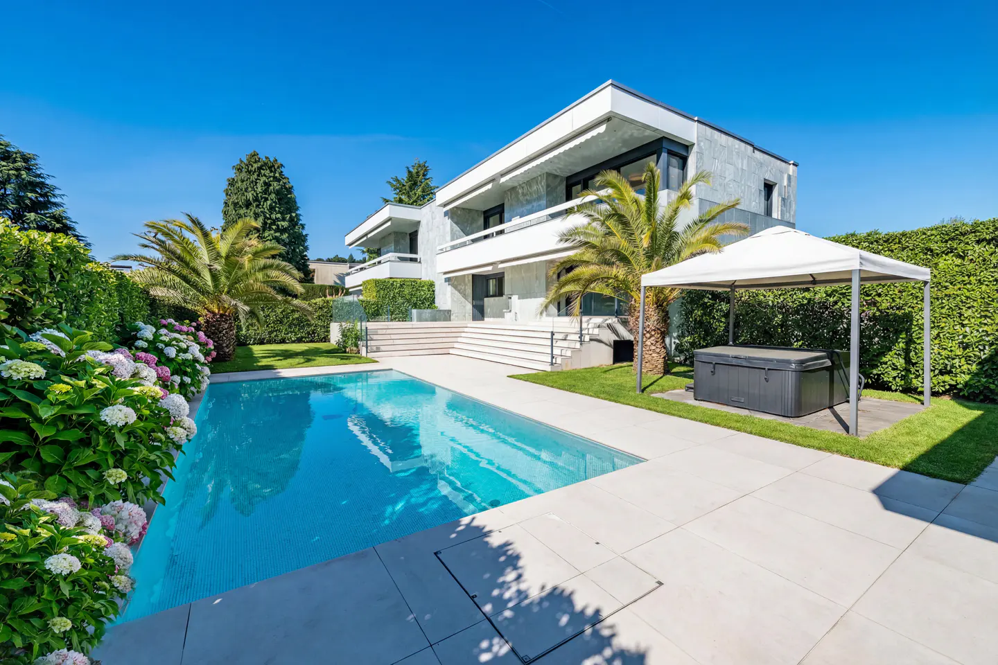 Modern two-story house with a pool, palm trees, and a hot tub under a white canopy on a sunny day.