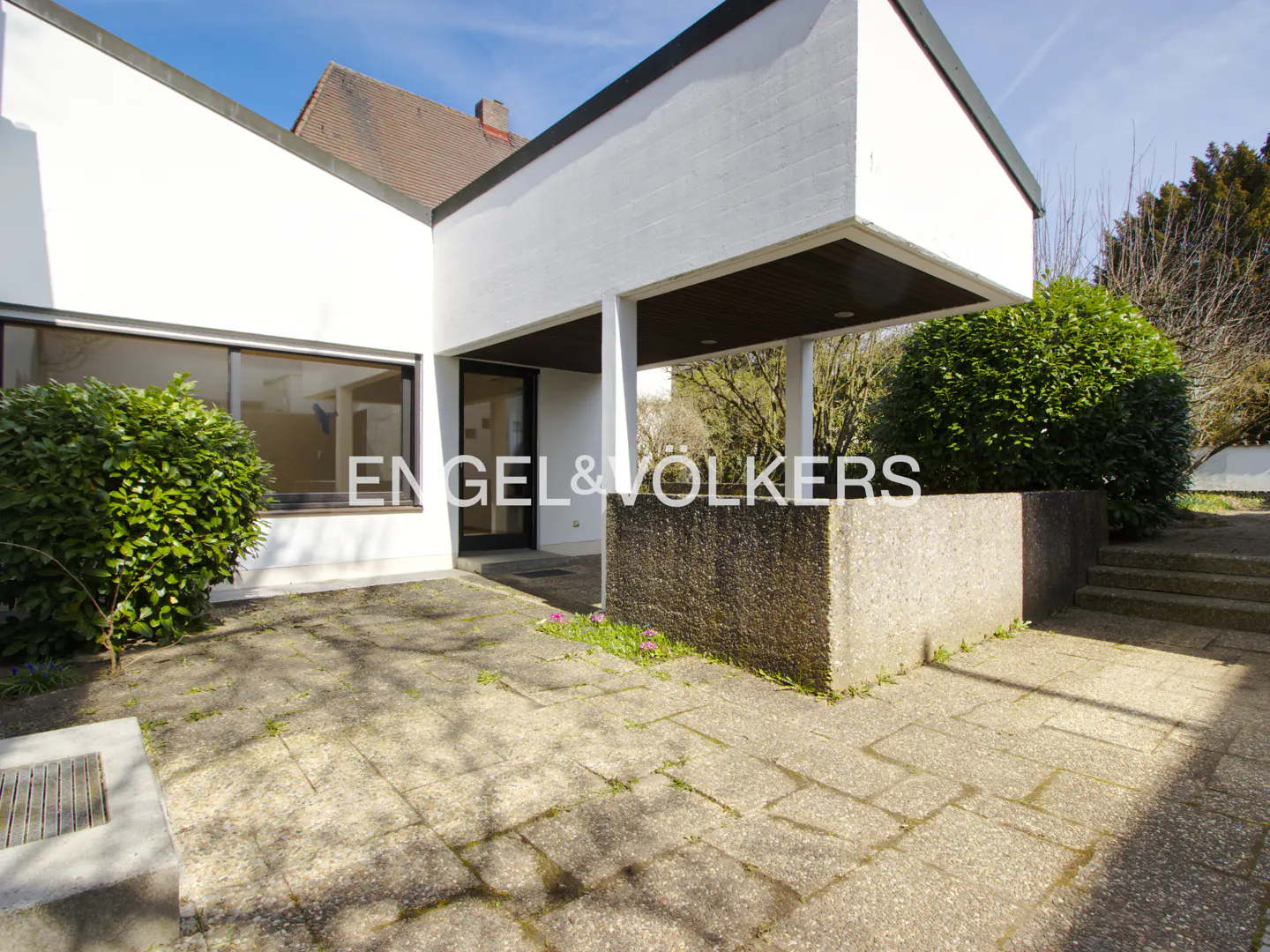 Exterior view of a modern white building with a covered entrance, stone patio, and green bushes. "Engel & Volkers" logo is visible.