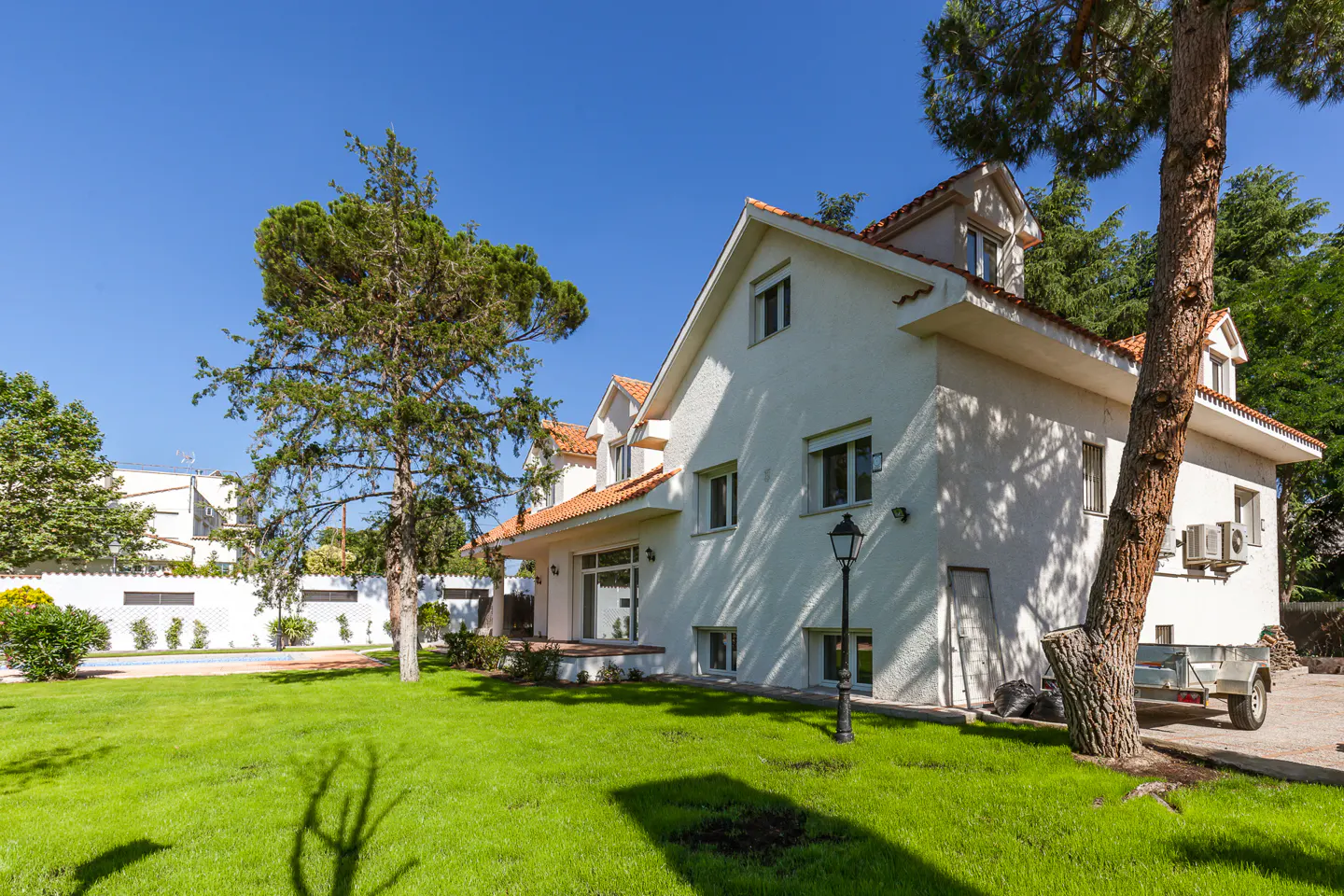 White two-story house with orange roof tiles, large green lawn, and tall pine trees under a clear blue sky.