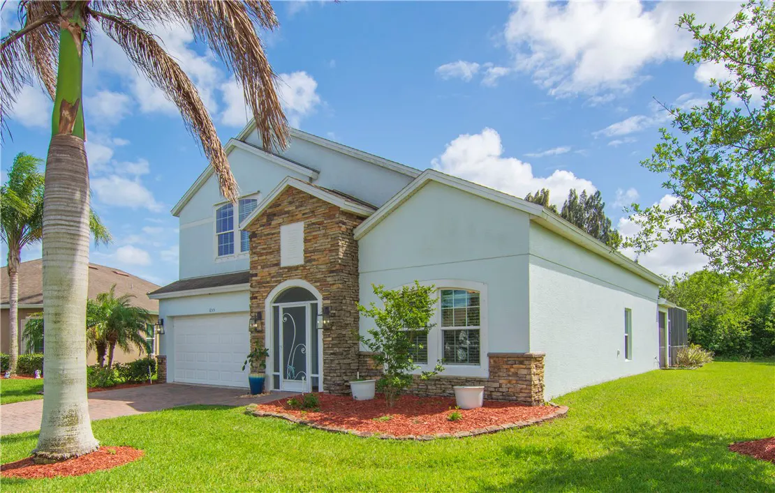 Two-story light blue house with stone facade, white trim, and green lawn under a blue sky with scattered clouds.