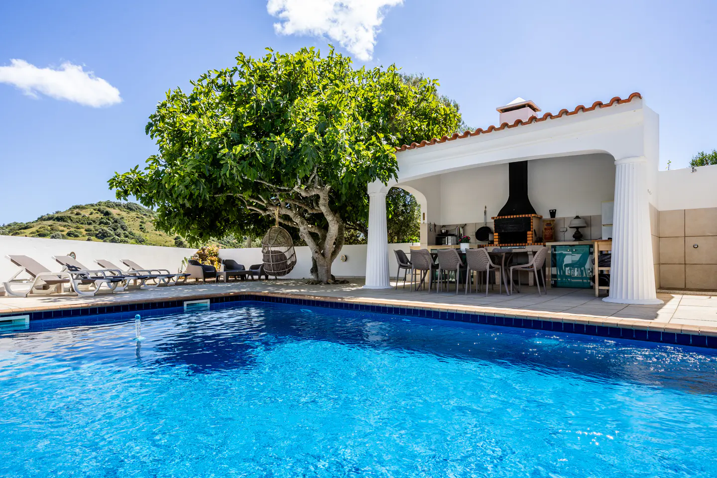 Outdoor pool with lounge chairs, a hanging chair under a large tree, and an outdoor kitchen with a dining table.
