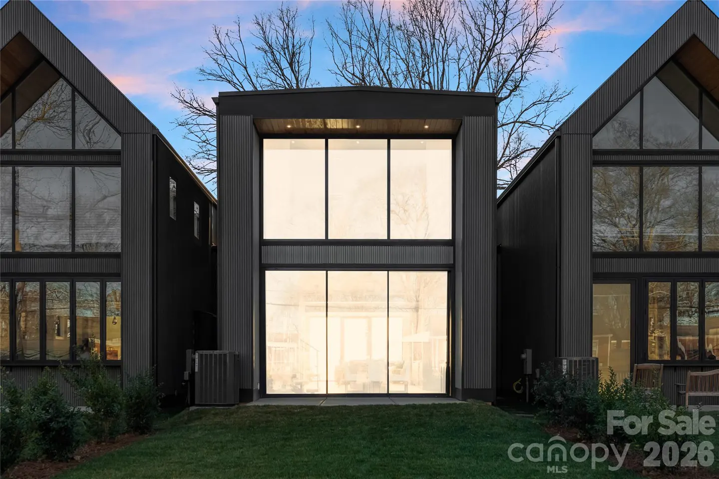 Modern black home exterior with large floor-to-ceiling windows, a grassy lawn, and a sunset sky in the background.