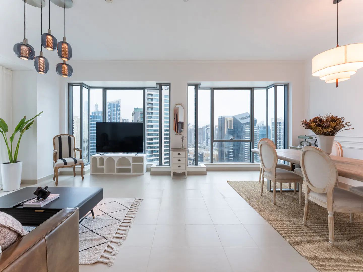Bright, modern apartment interior with city views. Living room with TV, dining table, and large windows. Neutral color palette.