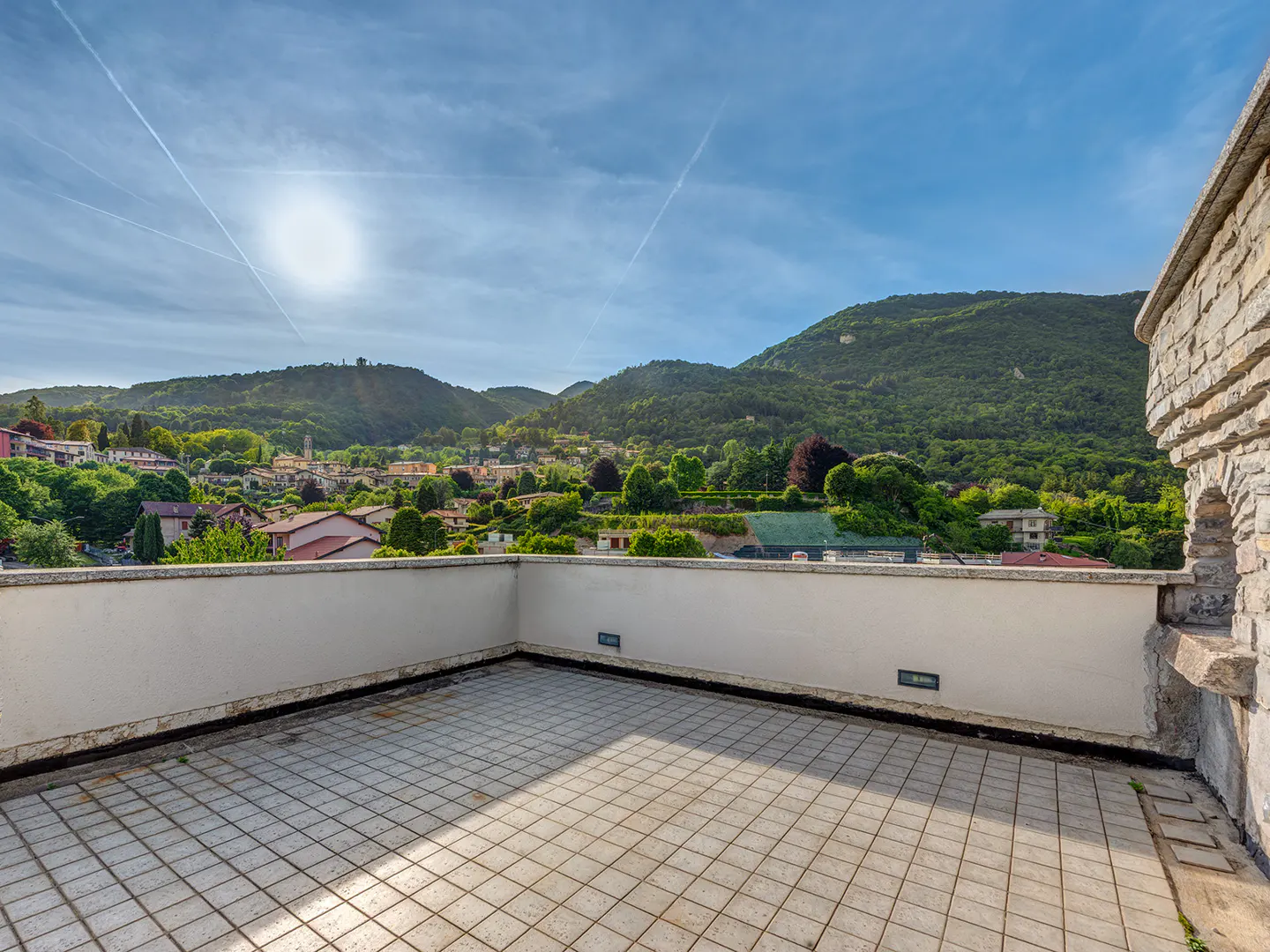 Spacious rooftop terrace with tiled floor, stone wall, and panoramic views of a lush green hillside town under a blue sky.