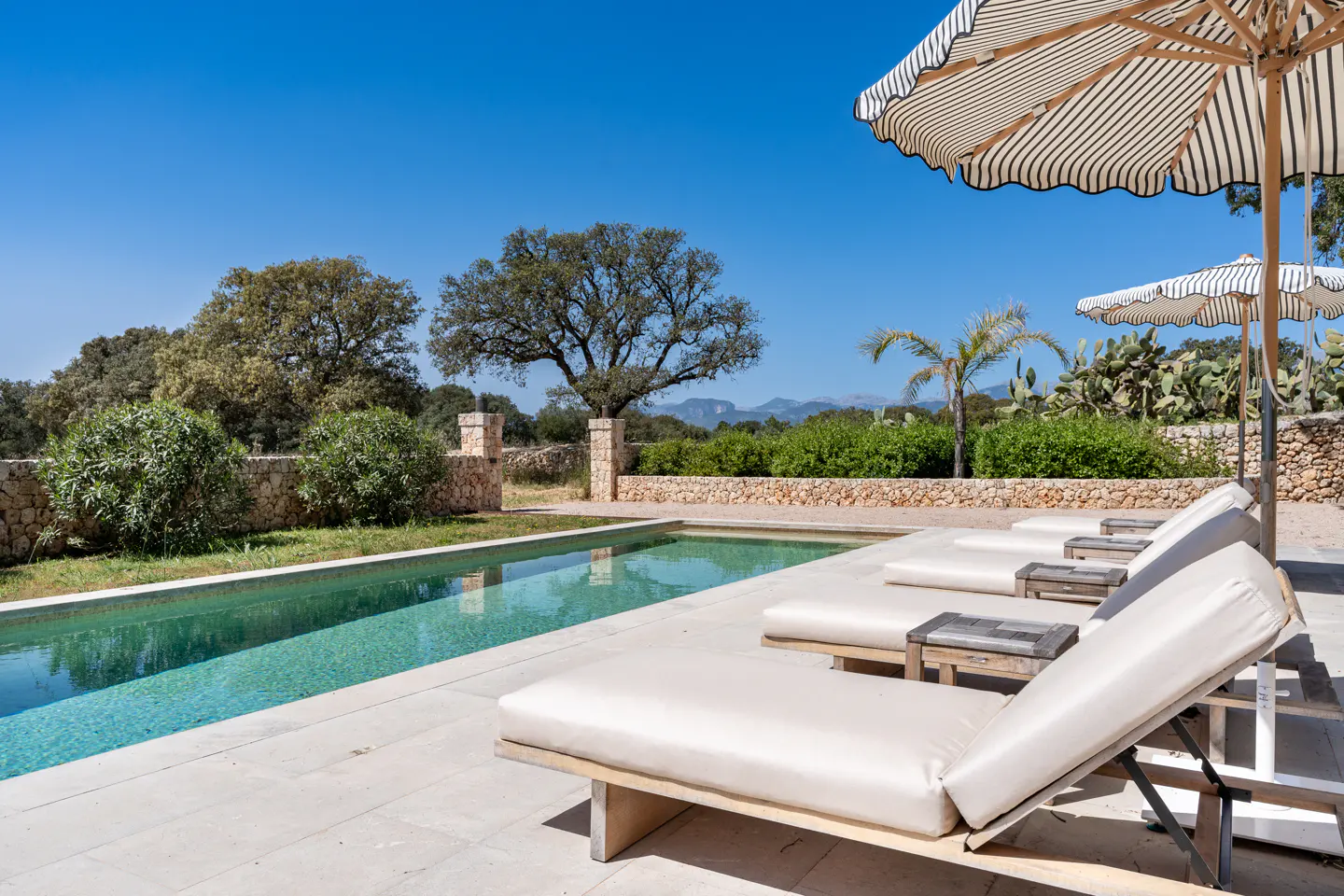Outdoor pool with lounge chairs and striped umbrellas. Stone wall and trees in the background under a clear blue sky.
