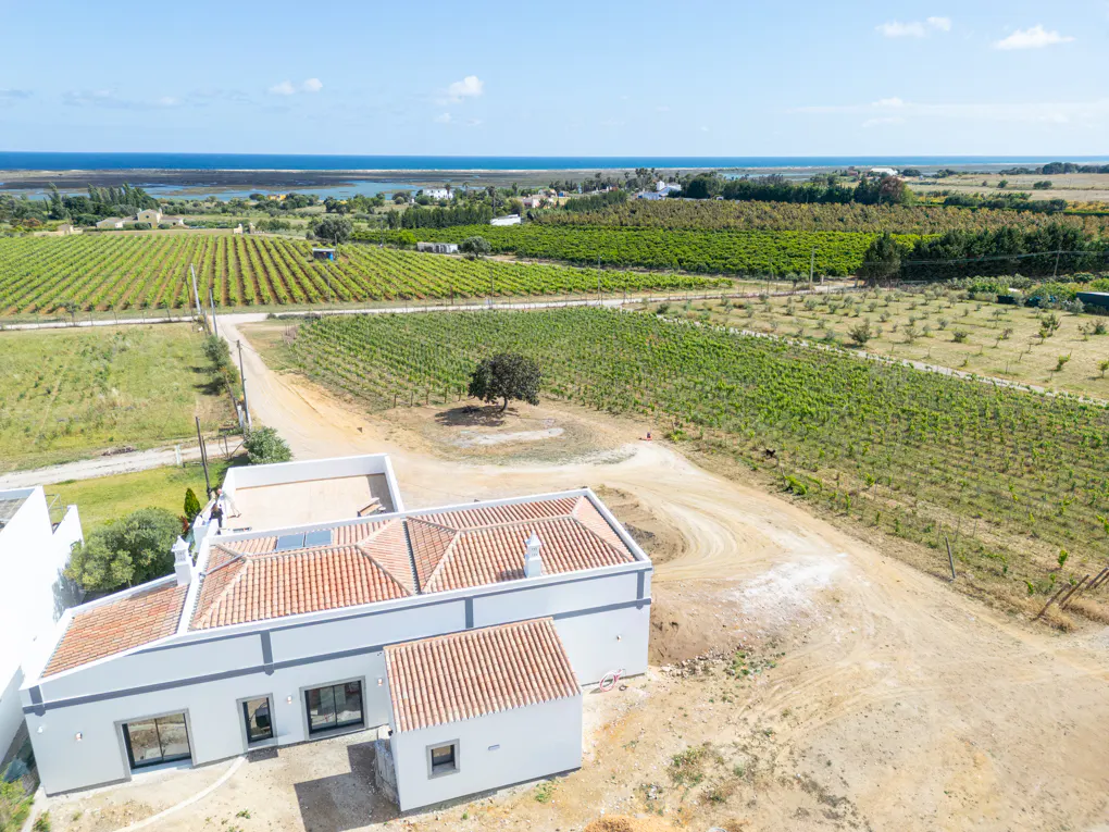 Aerial view of a white house with a red tile roof, surrounded by green vineyards and a blue ocean in the background.