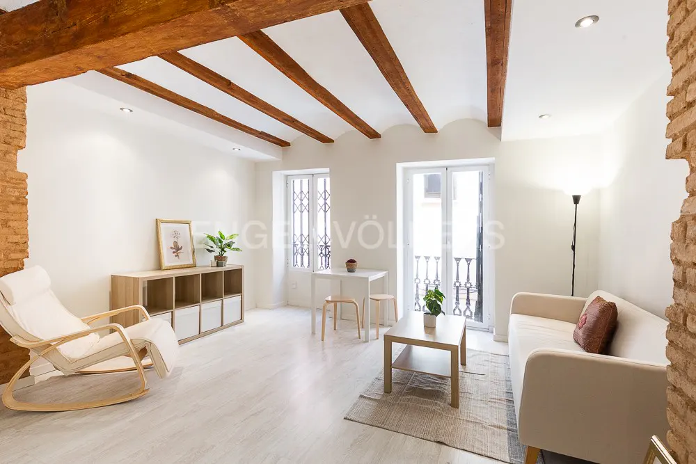 Bright living room with white walls, wooden beams, and exposed brick. Furniture includes a rocking chair, sofa, and small table.