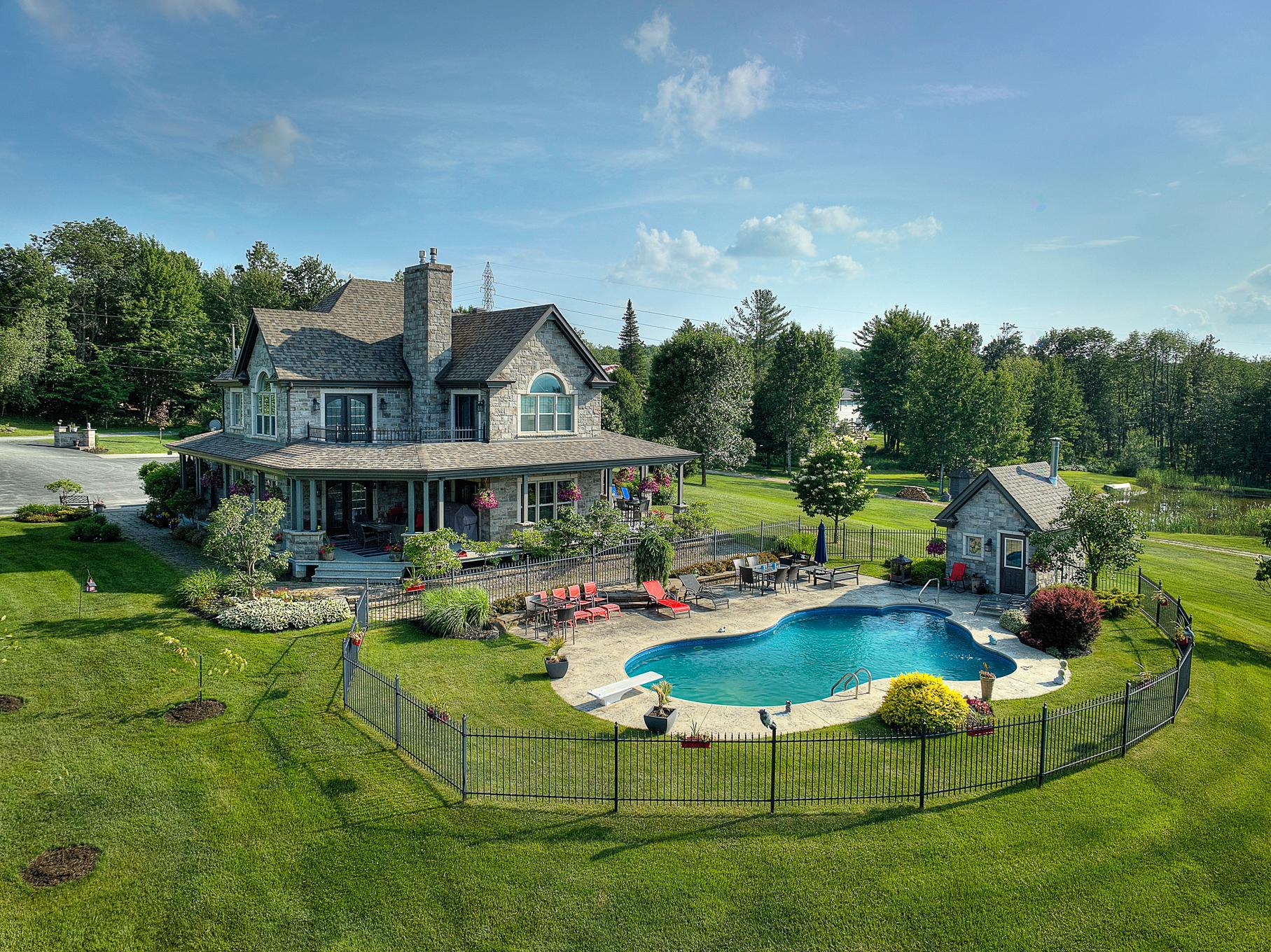 Aerial view of a large stone house with a pool, patio furniture, and a pool house on a green lawn.