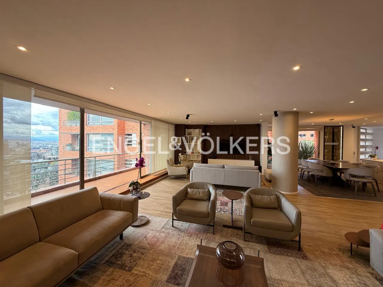 Wide shot of a modern living room with beige sofas, chairs, and a large window overlooking a city. Engel & Völkers logo visible.