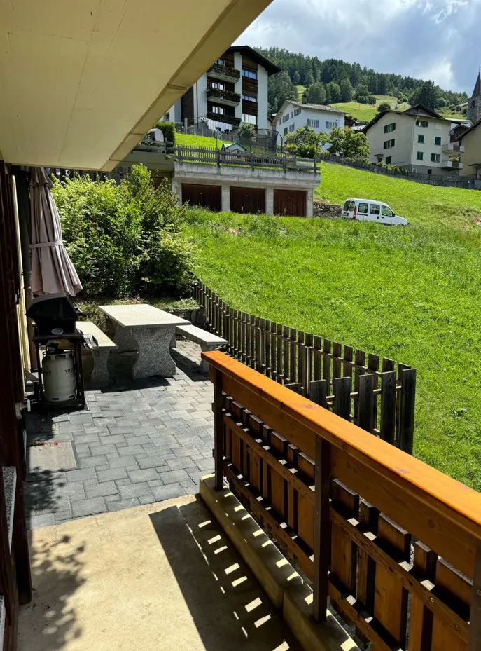 Outdoor patio with stone table, grill, and umbrella. A green lawn and houses are in the background.