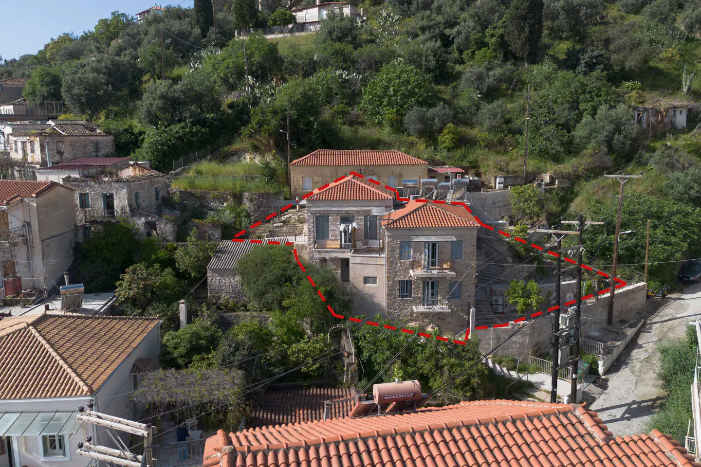 Two-story stone house with red tile roof, surrounded by trees and other buildings. Red dotted line marks property boundary.