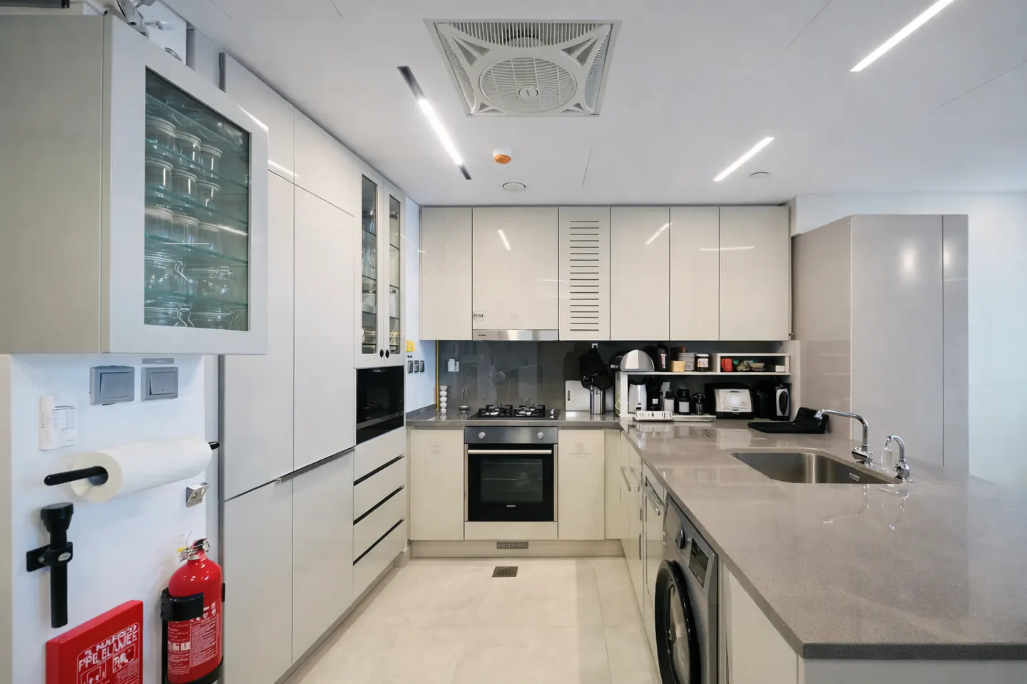 Bright, modern kitchen with white cabinets, gray countertops, and stainless steel appliances. A fire extinguisher is mounted on the wall.