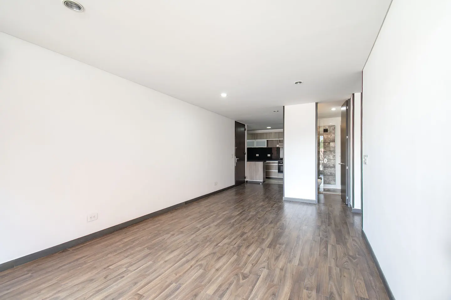 Bright, empty room with wood floors and white walls. A kitchen and bathroom are visible in the background.