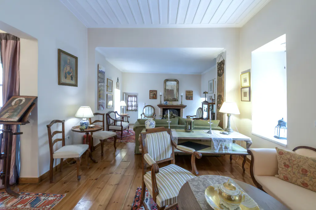 A bright, antique-filled living room with wooden floors, white walls, and a green sofa. Paintings and lamps decorate the space.