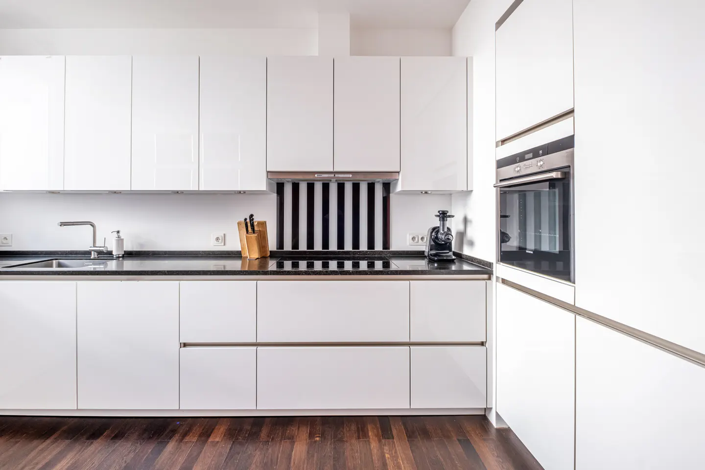 Modern kitchen with white cabinets, black countertop, and dark wood floors. Stainless steel oven and faucet are visible.