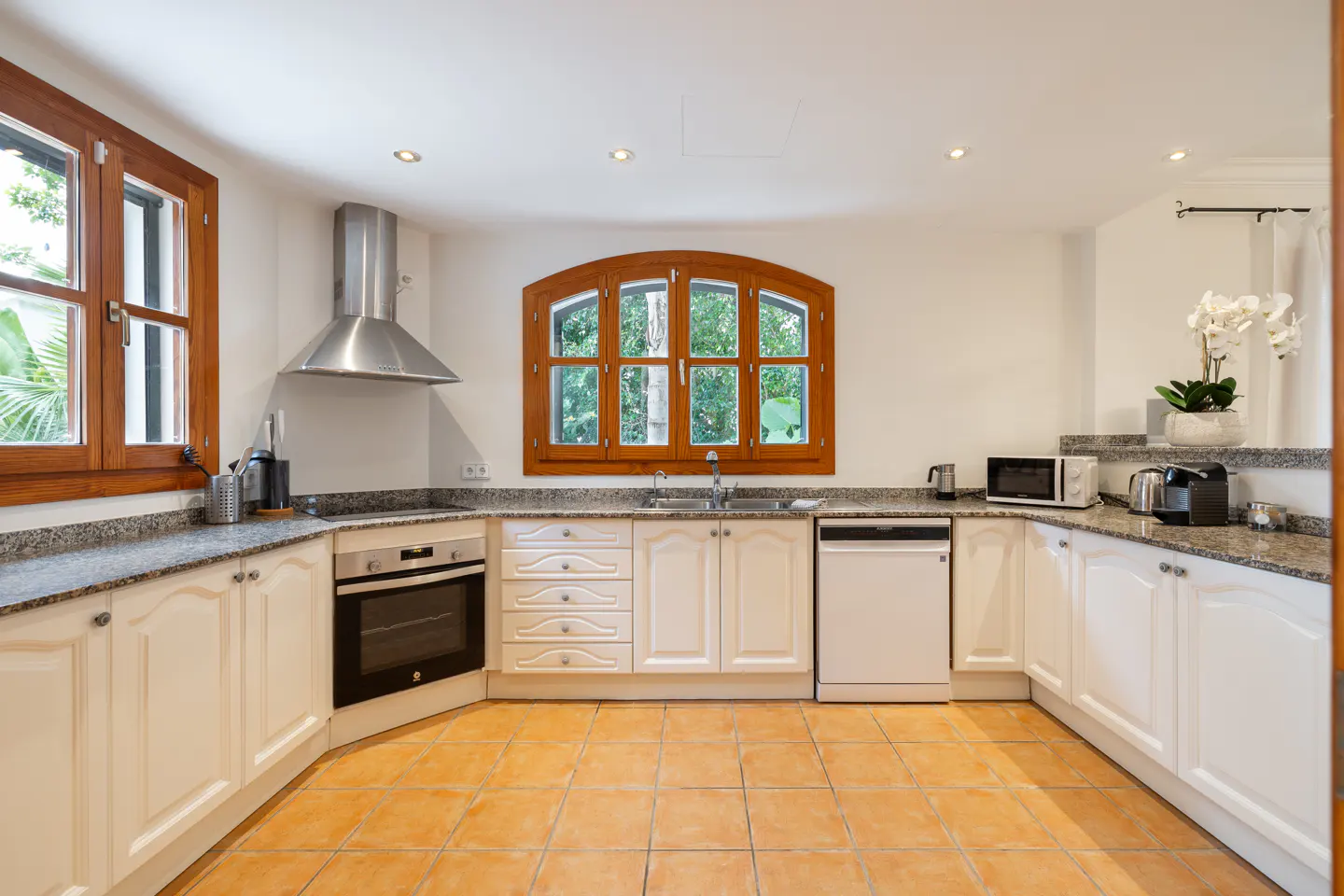 Bright kitchen with white cabinets, granite countertops, and terracotta tile floor. Wood-framed windows offer a view of lush greenery.