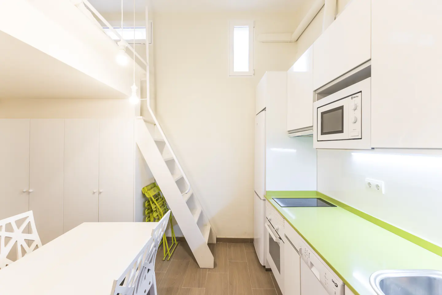 Bright, modern studio apartment with white cabinets, green countertops, and a loft bed accessed by a white ladder. A white table and chairs sit nearby.