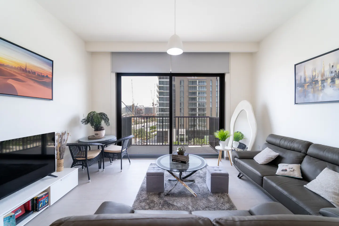 Bright living room with gray sofas, glass table, and balcony view of city buildings. Art on the walls adds color.