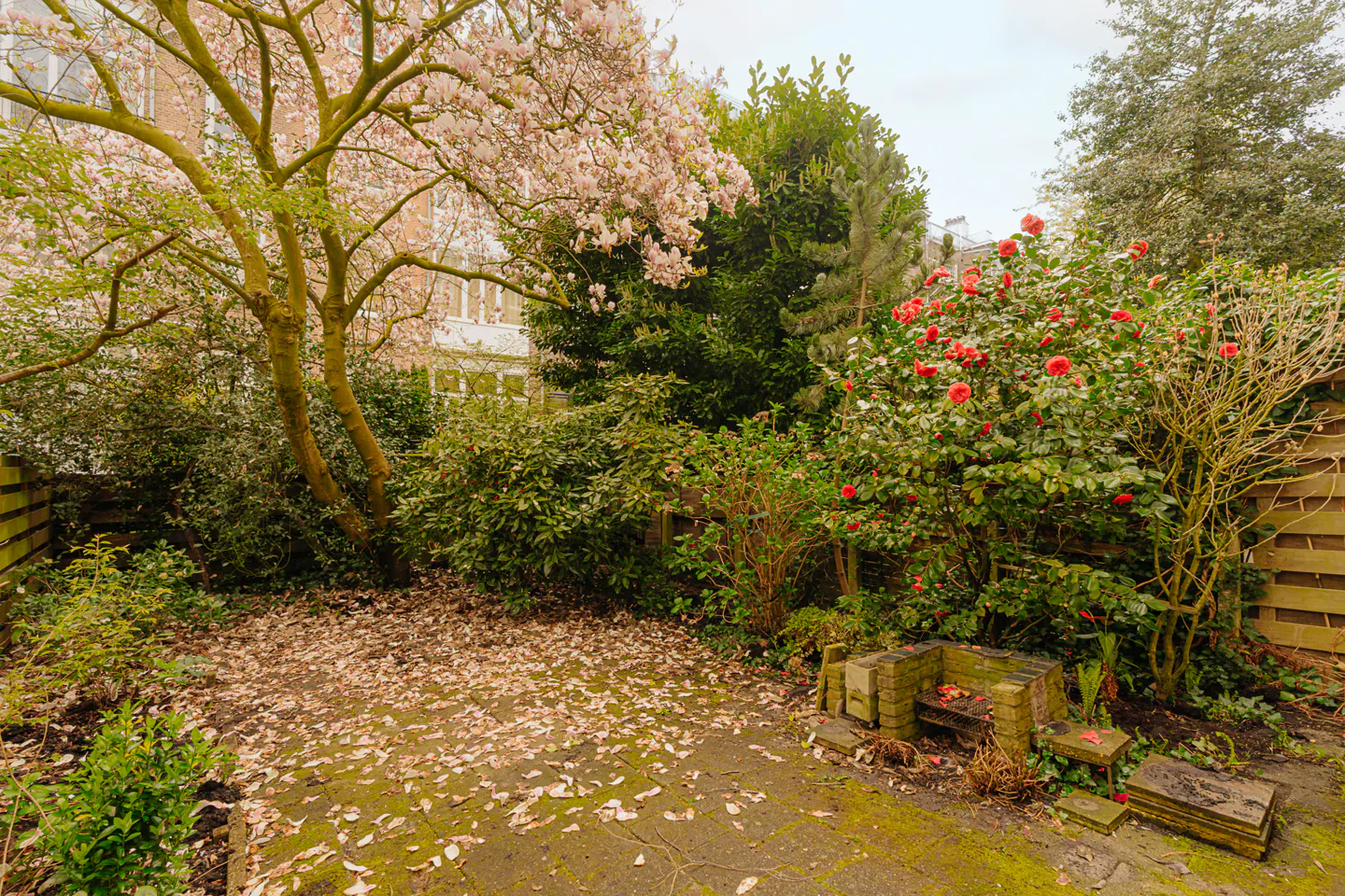A lush garden with pink blossoms, red flowers, and a stone patio covered in fallen leaves.