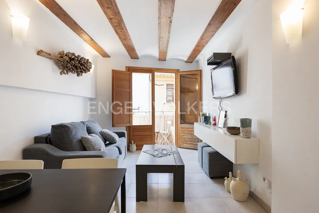Living room with gray sofa, black table, and white walls. Wooden beams on the ceiling and a balcony with open doors in the background.