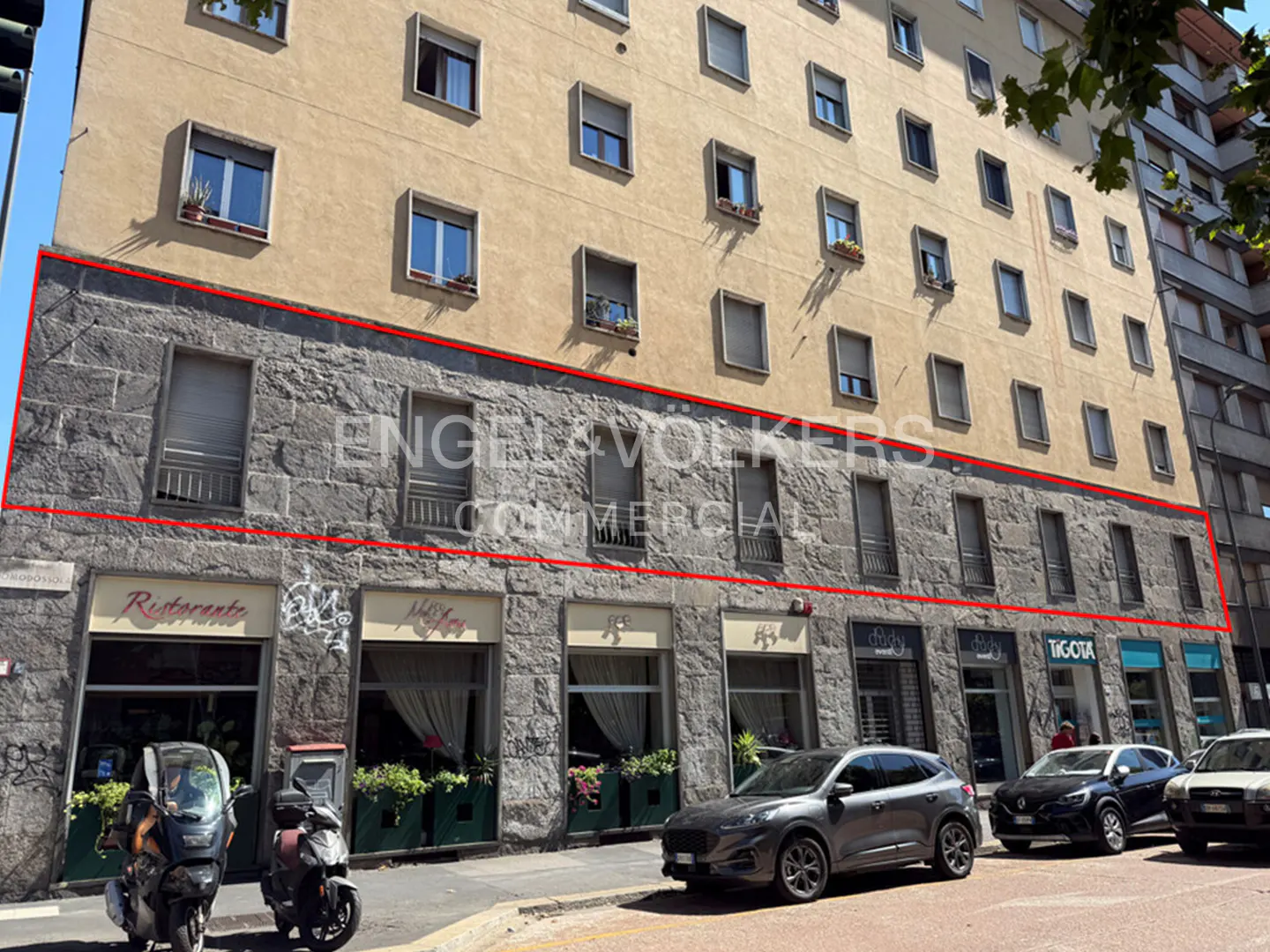 Commercial building with stone facade and red outline, street view. Restaurants and shops on the ground floor, cars parked in front.
