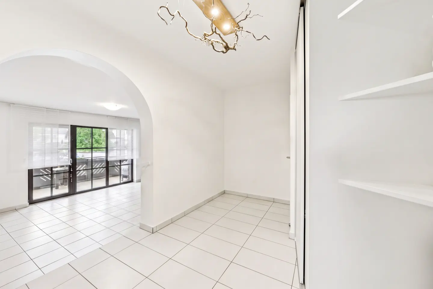 Bright, all-white hallway with tile floor, arched doorway to a balcony, and a unique branch-like ceiling light fixture.