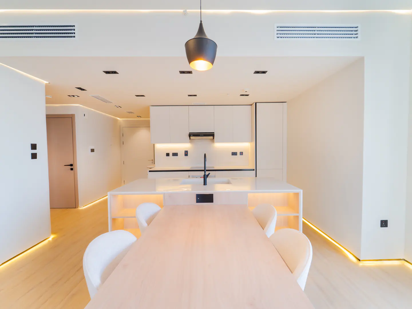 Bright, modern kitchen with white cabinets, island, and dining table. A black pendant light hangs above the island. LED strip lighting accents the floor and cabinets.