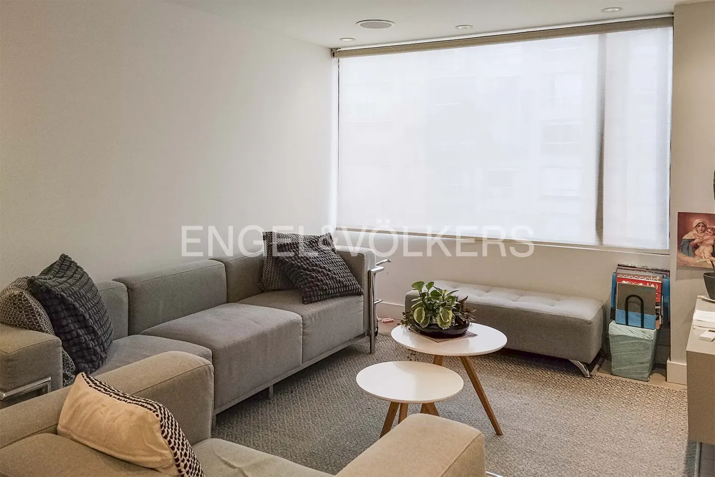 Modern living room with a grey sectional sofa, two small round white coffee tables, and a large window with white blinds.