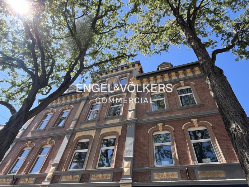 Low-angle view of a historic red brick building with ornate trim, framed by large trees against a bright blue sky.