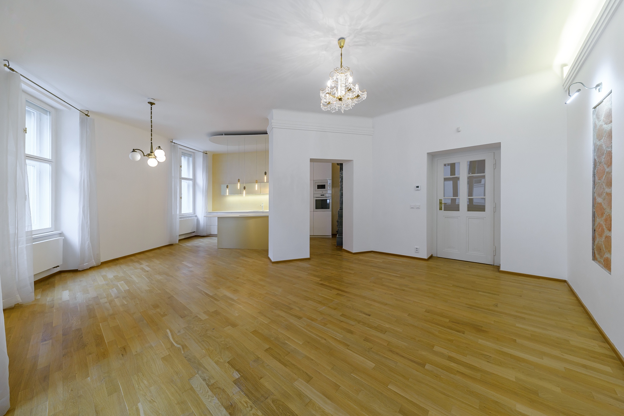 Spacious, bright living room with light wood floors, white walls, crystal chandelier, and a view into a modern kitchen. [AI-generated text]