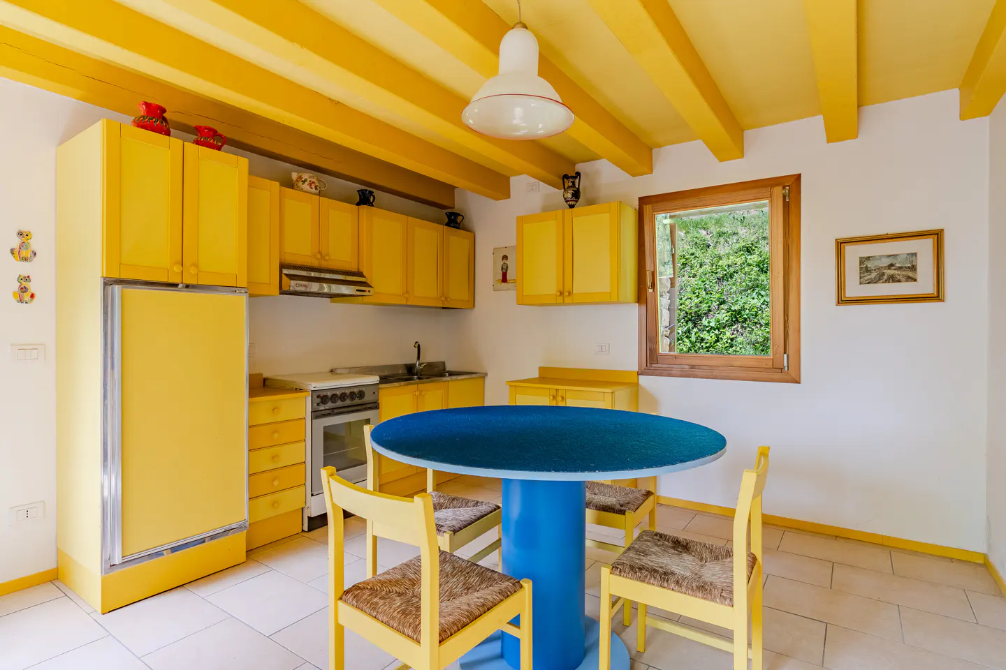A bright kitchen with yellow cabinets, ceiling beams, and chairs. A blue round table sits in the center. A window overlooks green foliage.