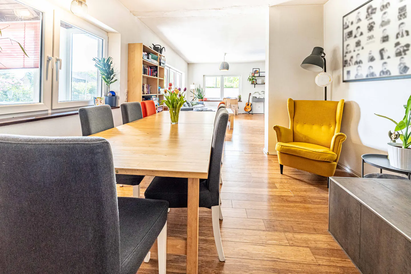 A bright, open-concept dining and living room with a long wooden table, gray chairs, and a yellow armchair.