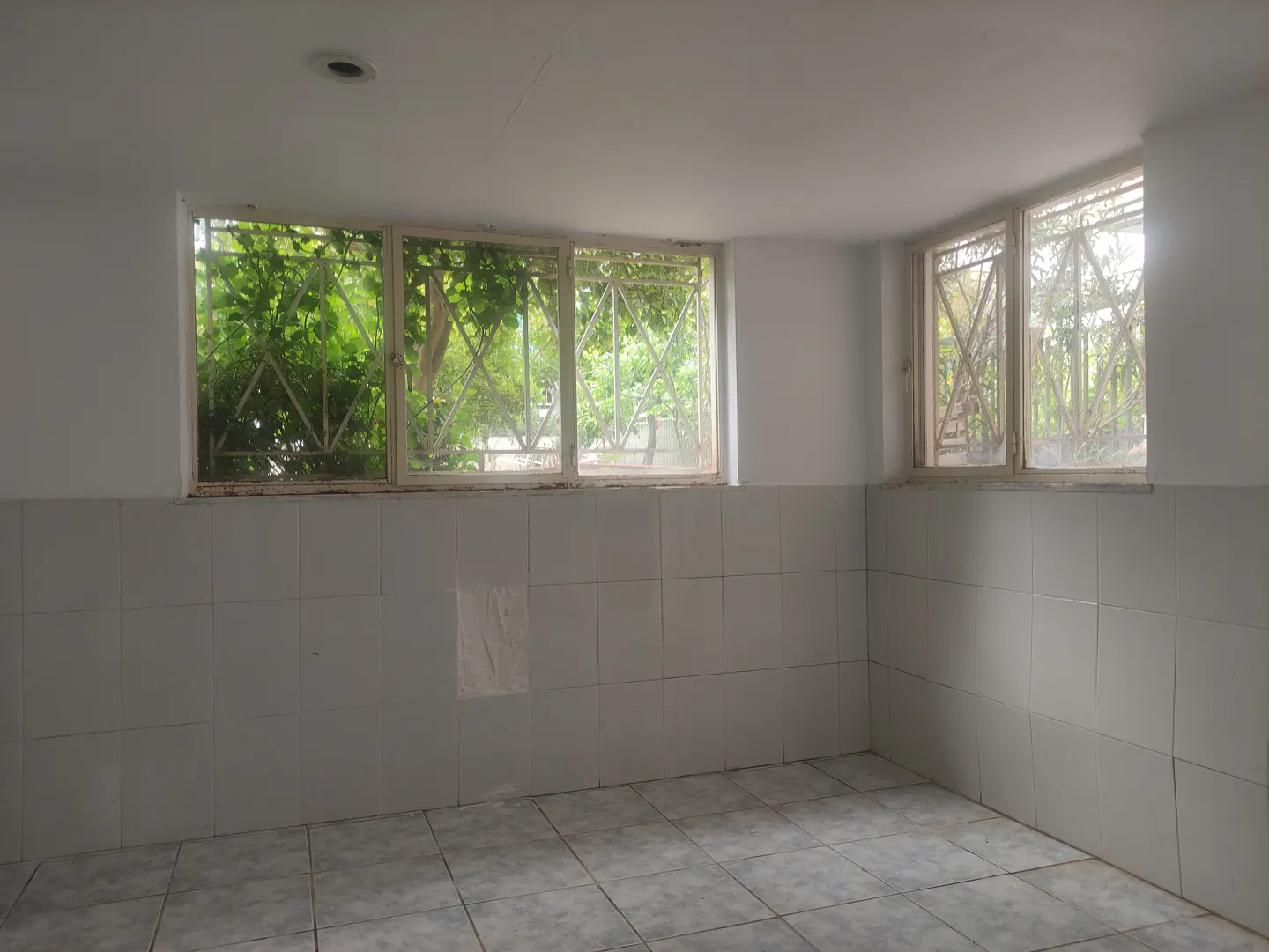 Empty room with white walls, tiled halfway up. Two barred windows show green foliage outside. Tiled floor.