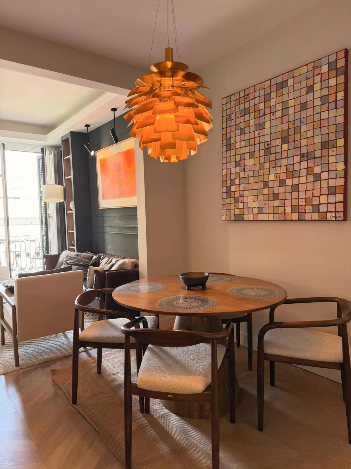 A dining room with a round wooden table, chairs, and a unique orange pendant light. A colorful, grid-patterned artwork hangs on the wall.