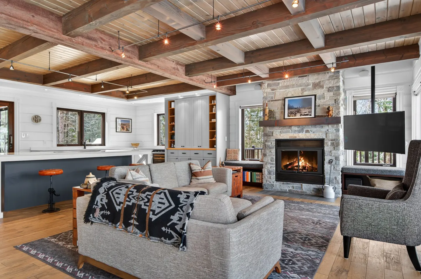 Open-concept living room with a stone fireplace, gray sofas, and a wooden-beamed ceiling. A black TV hangs near a window.
