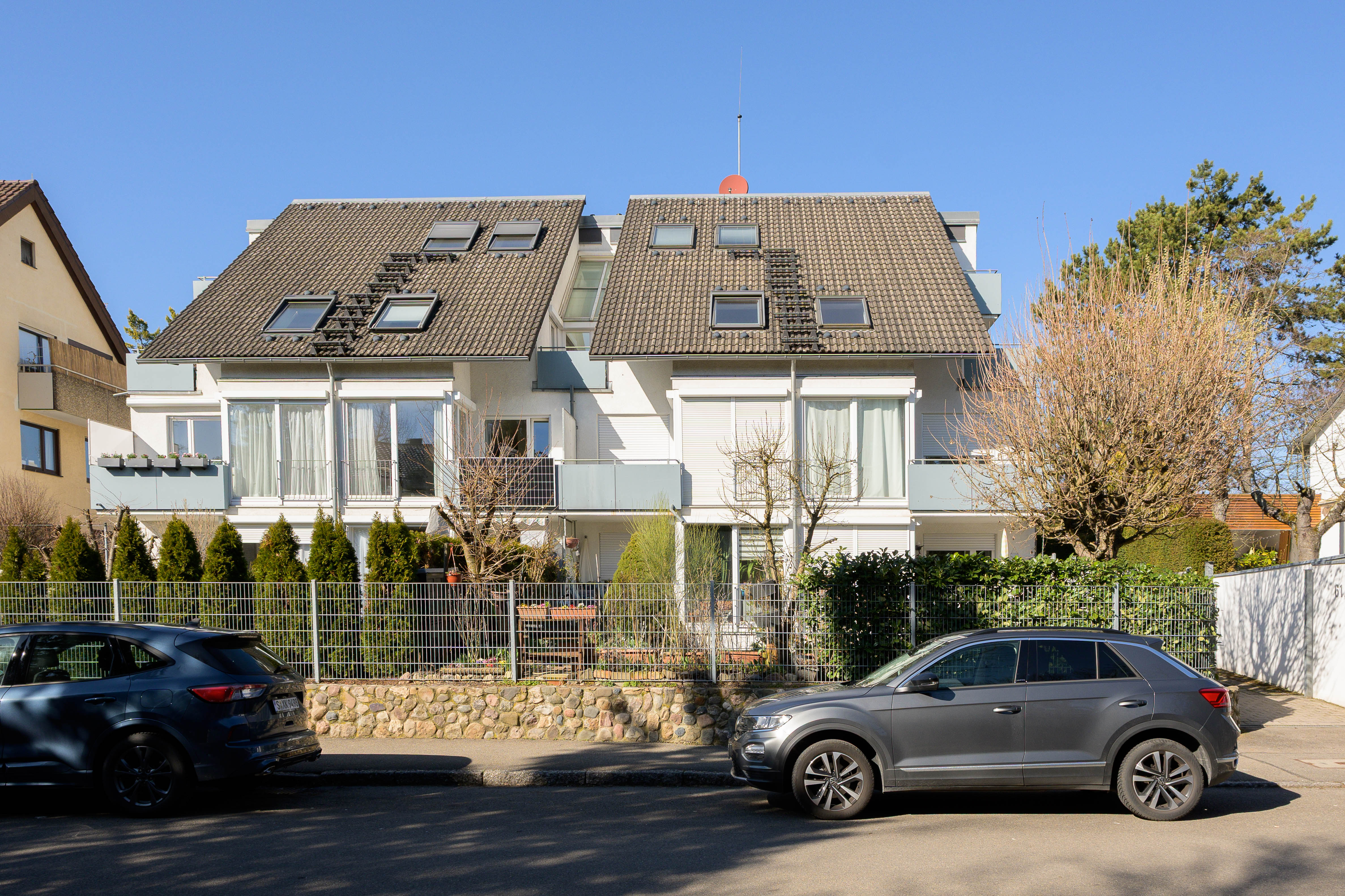 A modern white two-story townhouse with a tiled roof, fenced garden, and two cars parked on the street in front.