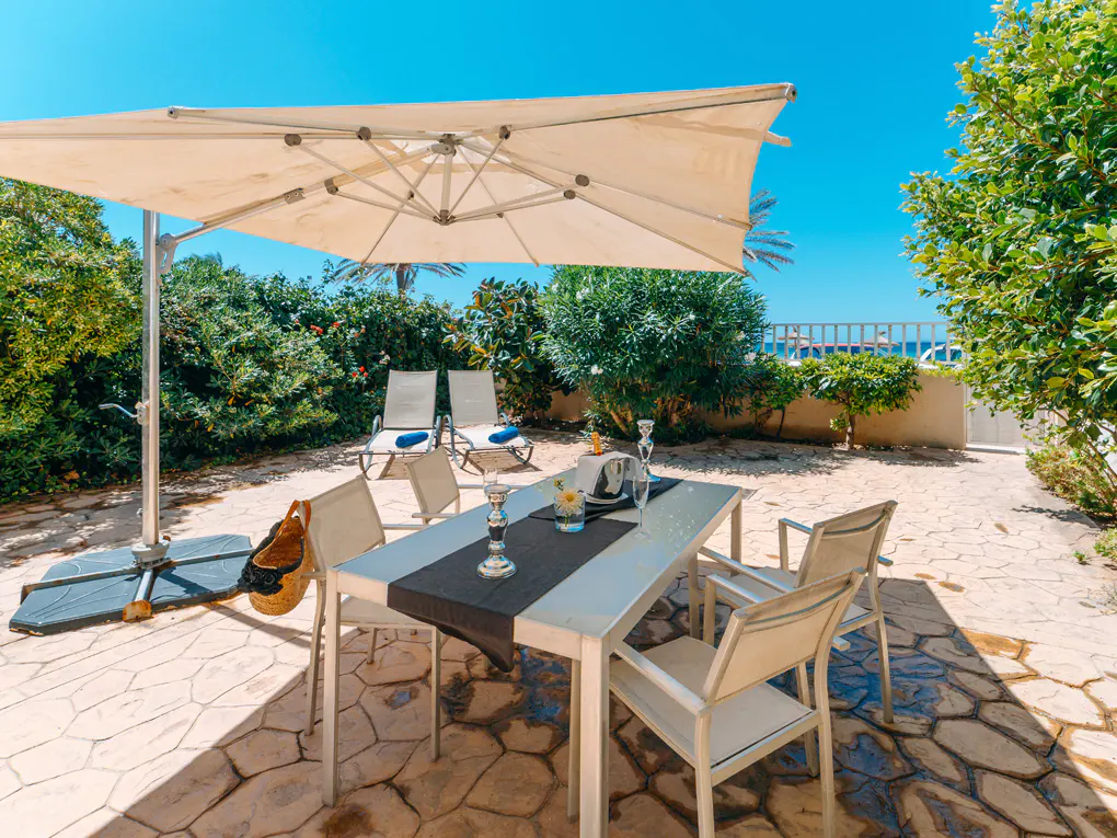 Outdoor patio with a white table set for dining, chairs, umbrella, and lounge chairs under a clear blue sky.