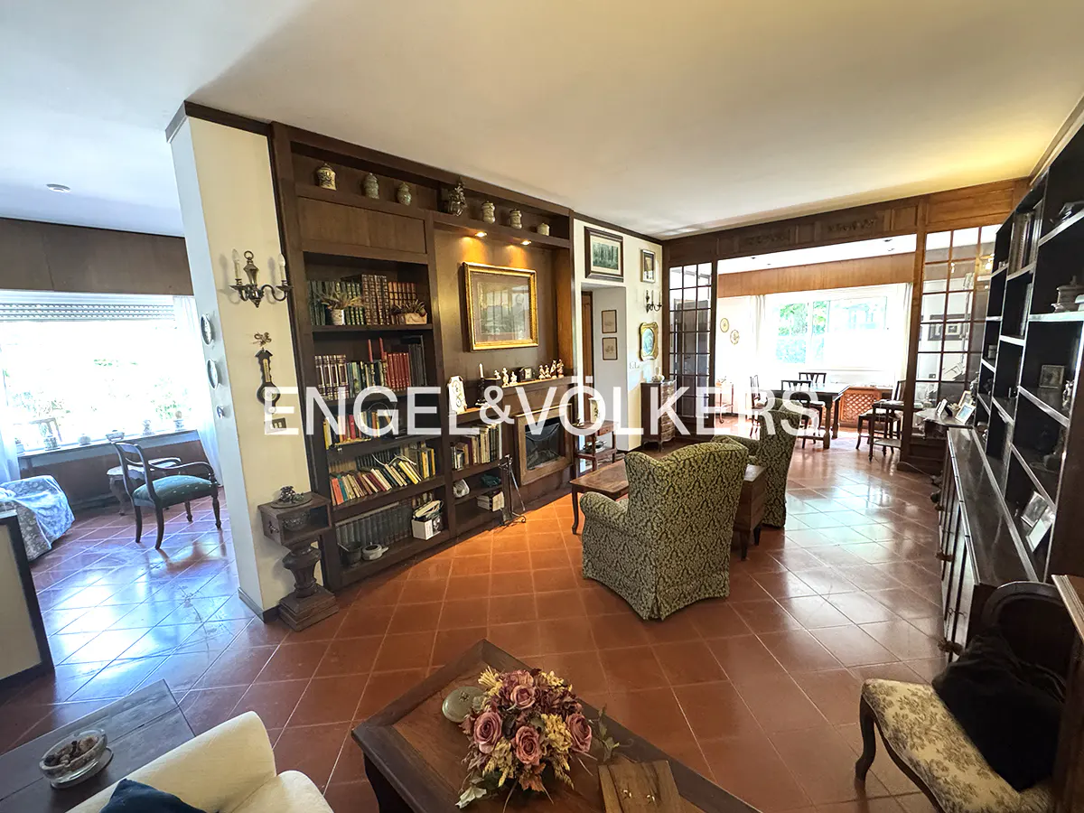 A wide shot of a living room with a dark wood bookcase, patterned chairs, and terracotta tile flooring.