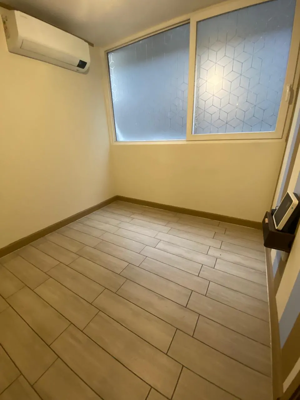A bright, empty room with wood-look tile flooring, a large window with frosted glass, and an air conditioning unit.