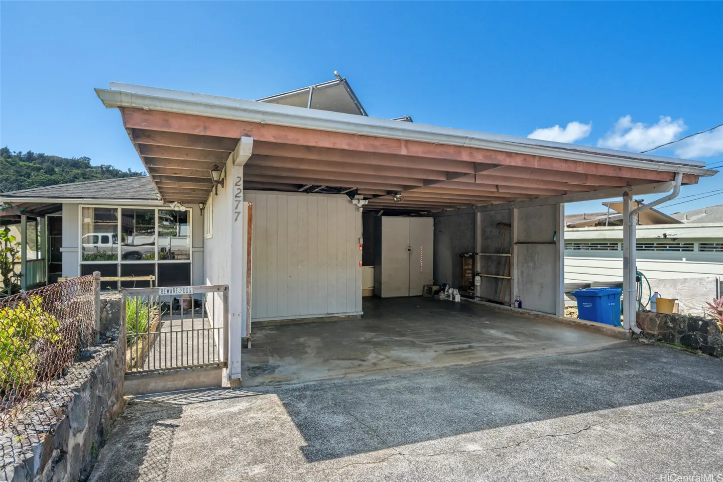 Exterior view of a house with a carport. The carport has a wooden roof and concrete floor. A blue trash bin sits on the side.