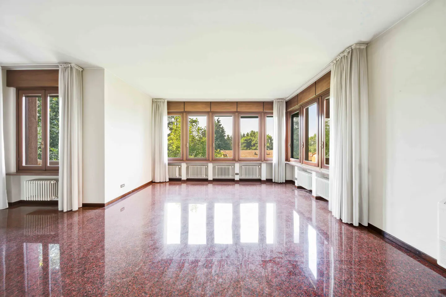Empty room with polished red granite floors, white walls, and large windows with wooden frames and white curtains.