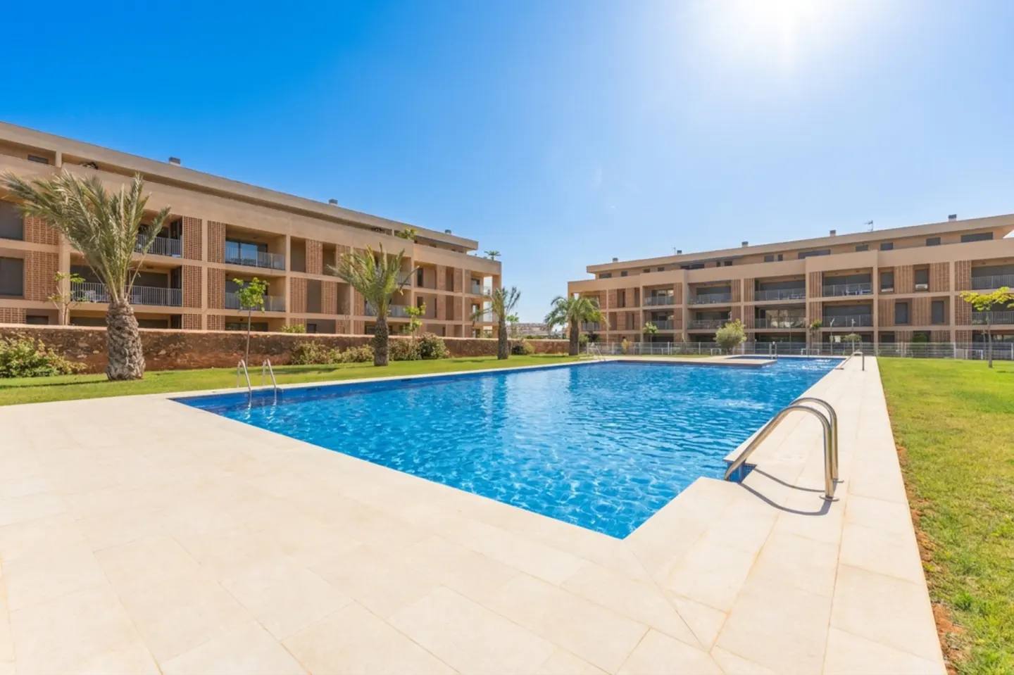 Outdoor pool with blue water and beige tile surround. Two apartment buildings with balconies flank the pool, with palm trees and green grass.